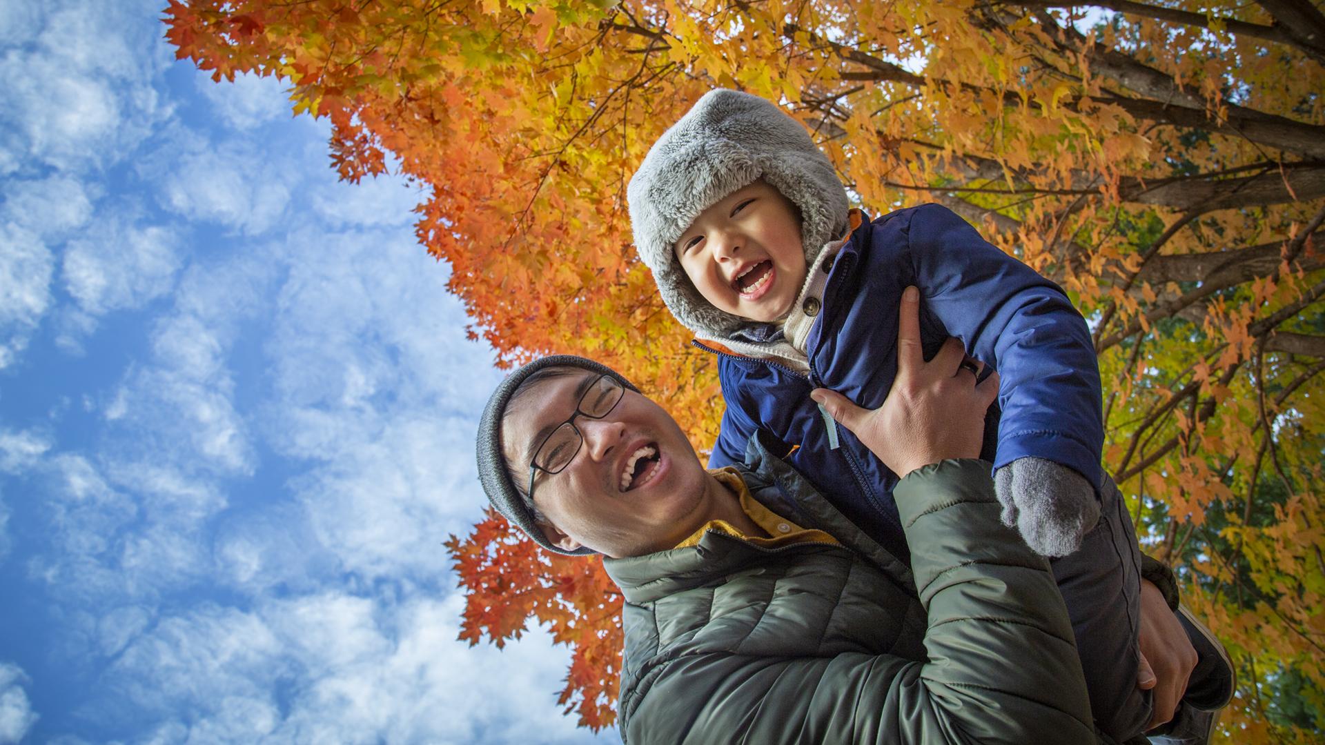 All of Us Hmong father holding young son outdoors, both smiling at camera in front of maple tree with orange leaves